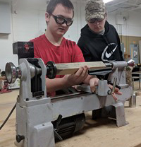 Two students examines a piece of wood on a lathe.