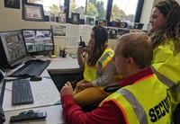 three career and technical education students wearing security vests use a radio and look at security monitors