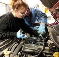 Two students wearing protective googles and gloves reach inside the engine of a car