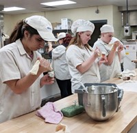 Three students in baking smocks and hats squeeze pastry bags to create frosting roses.