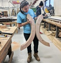 In a carpentry classroom, a student holds up for display two large curved wooden table legs she made.