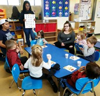 In a colorful classroom, six preschool children sit around a table. One student in the Child and Family Development program holds a chart with alphabet letters while two other high school students look on. 