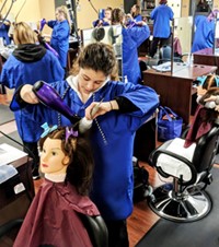 A student in a smock uses a blow dryer and brush on a mannequin head that is supported on a salon chair, while several other students also working on mannequins are visible in the classroom background.