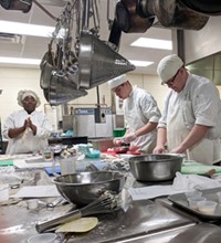 Three students in chef's hats and aprons prepare food at a large steel prep table, with many cooking tools and pots hanging overhead and on the table.