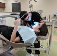 Two dental assisting students work together. One is fully reclined in a dental chair and holding a mirror. The other is wearing gloves, a mask and uniform, and is examining the first student's teeth. 