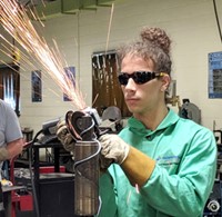 A student in full safety gear uses a hand held power tool that is creating a shower of sparks while sanding metal. 