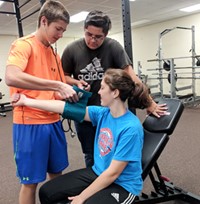 A student sitting on exercise equipment has her blood pressure taken by another student, while a third student looks on.