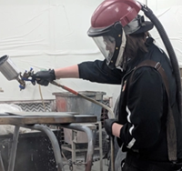 A student wearing a respirator helmet uses a professional sprayer to paint a metal panel.