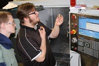 A student examines a metal part while another looks on, with a CNC lathe behind them.