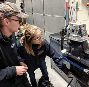 In a classroom a student wearing safety glasses make an adjustment to a hose that is attached to a boat motor.