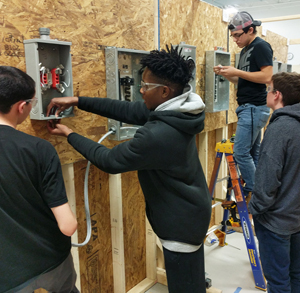 Four students work in an electricity classroom, working in pairs to wire junction boxes mounted to a wall. 